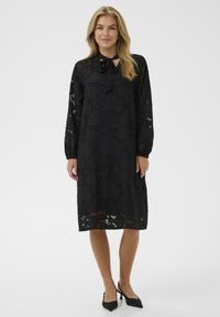Rochie neagră până la genunchi, cu mâneci lungi, din material texturat, detaliu cu fundă la gât și accente transparente. Purtată cu pantofi cu toc ascuțit negri.