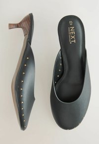 Mules de tacón de cuero negro con textura suave, con tachuelas doradas a lo largo de los lados y un tacón de madera. Marca etiquetada en la plantilla.