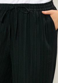 Pantaloni neri in tessuto testurizzato con cintura elastica e coulisse, tasca interna posizionata a mano sul lato destro.