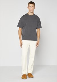 T-shirt gris surdimensionné à manches courtes, associé à un pantalon couleur crème et des chaussures ouvertes marron, se tenant sur un fond uni.