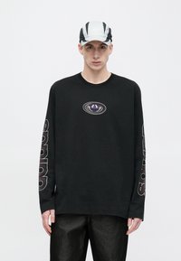 Juoda ilgomis rankovėmis marškinėliai su violetiniu ir baltu logotipu centre bei baltu užrašu ant rankovių. Vyras dėvi dviejų spalvų kepurę.