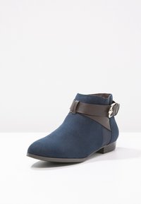 Marinblå mocka ankelsko med ett brunt läderrem med spänne. Snygg design, spetsig tå och låg klack. Strukturerad yta.