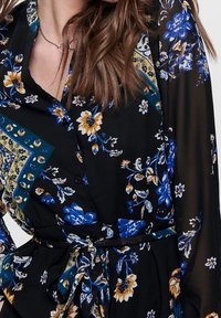 Robe noire avec motif floral comprenant des fleurs bleues et jaunes ; manches longues, patte de boutonnage à l'avant et ceinture à nouer à la taille. Tissu transparent.