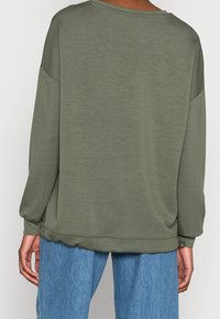 Osoba ubrana w oliwkową długą bluzę i niebieskie dżinsy, pokazana od tyłu na jednolitym tle.