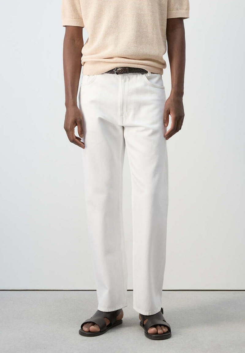 Personne portant un pull beige à manches courtes, un pantalon blanc droit avec une ceinture noire, et des sandales noires à bouts ouverts sur un sol gris.
