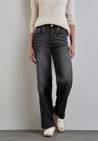 Jeans de denim negro de pierna ancha con un ligero desgaste y cinco bolsillos, combinados con un suéter crema y zapatillas blancas. Acentos de costura simples.