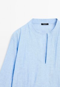 Chemise en lin bleu clair avec un col rond, un plastron central et des manches longues, agrémentée d'une texture subtile et de détails de couture.