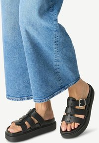 Black leather sandals with a thick sole, featuring crossed straps and a side buckle. Paired with loose, light blue denim pants.