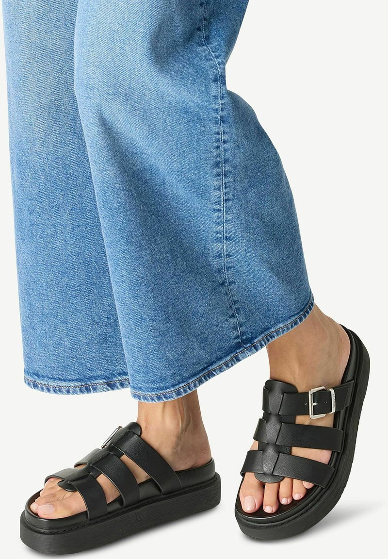 Black leather sandals with a thick sole, featuring crossed straps and a side buckle. Paired with loose, light blue denim pants.