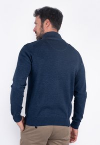 Pull azul marino de un suave tejido de punto con un cuello alto, puños y dobladillo acanalados. Presenta una yugo redondeado en la parte posterior y un sutil detalle de logo.