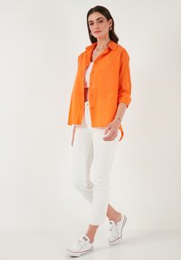 Oranžová košile na knoflíky, bílý crop top, bílé džíny a bílé tenisky. Košile má dlouhé rukávy, které jsou vyhrnuté, a volný střih.