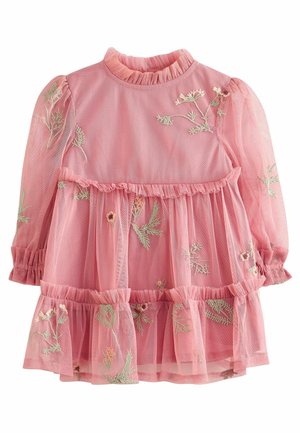 Bambino con vestito rosa ricamato, calze bianche e scarpe grigie tiene foglie verdi in piedi su pavimento di legno con tenda floreale sullo sfondo.