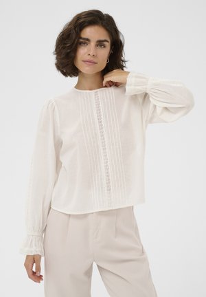 Femme aux cheveux courts et bouclés portant un chemisier blanc à manches longues avec des détails en dentelle et un pantalon beige taille haute, devant un fond uni.