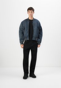 Giacca bomber navy con texture trapuntata, chiusura con zip, polsini a coste, abbinata a pantaloni neri e scarpe nere su uno sfondo bianco.