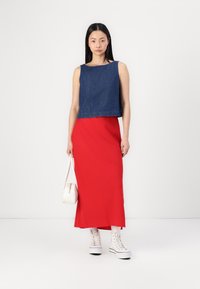 Denim crop top met een vierkante halslijn, gecombineerd met een felrode maxi rok. Model houdt een kleine witte handtas vast en draagt witte sneakers.