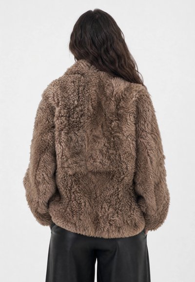 Personne aux longs cheveux foncés portant une veste ample en fausse fourrure marron et un pantalon noir, vue de dos sur un fond clair uni.