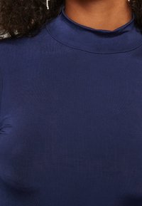 Námořnická modrá blůza s vysokým límcem vyrobená z hladkého, elastického materiálu. Má přiléhavý střih s jemným leskem a minimem švů.