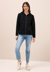 Felpa nera con zip, tasca a marsupio e cappuccio regolabile con coulisse, abbinata a jeans azzurri chiari e sneakers bianche.