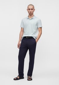 Jeune homme debout portant un polo gris clair, un pantalon bleu marine foncé avec ceinture noire et des sandales noires sur un fond clair uni.