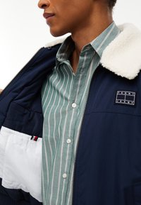 Homme portant une chemise à rayures vertes et blanches à boutons sous une veste bleu marine avec col en polaire blanche et écusson logo Tommy Jeans.