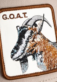 Embroidered patch featuring a goat head in brown, black, and white, with "G.O.A.T." text above on a light background. Brown border.