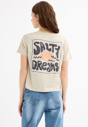Camiseta de algodón de color beige claro con un diseño corto. Presenta un gran gráfico que dice "SALTY DREAMS" con motivos de sol y olas en la parte posterior.