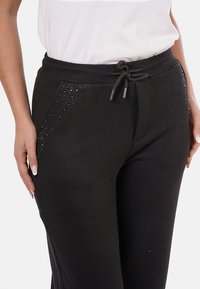 Donna che indossa pantaloni neri con vita elastica e piccole decorazioni lungo i bordi delle tasche, abbinati a una camicia bianca infilata dentro.