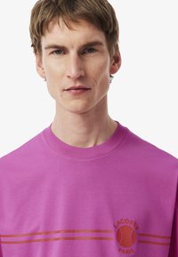 T-shirt in cotone viola con colletto rotondo a coste, righe orizzontali e stampa grafica con un motivo tennistico e testo "LACOSTE PARIS".