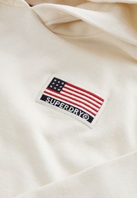 Krämfärgad huva med en broderad amerikansk flagga i röd, vit och blå färg samt en Superdry-logotyp i svart. Mjuk tygstruktur.