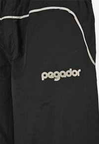 Črne hlače iz lahkega materiala, ki imajo bel trak ob žepih in blagovno znamko "pegador" v svetli barvi na strani.