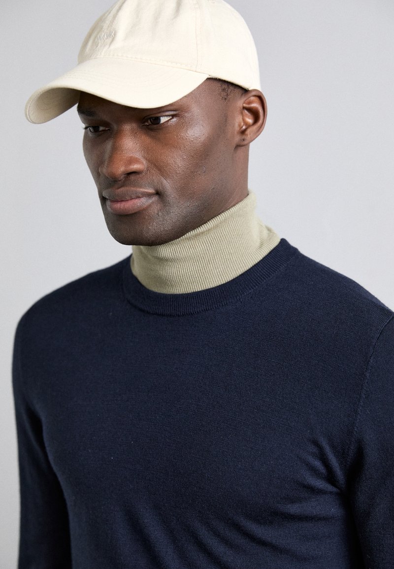 Suéter de punto azul marino con un cuello de tortuga acanalado en beige claro y una gorra de béisbol beige claro. Primer plano de la textura y el ajuste del tejido.