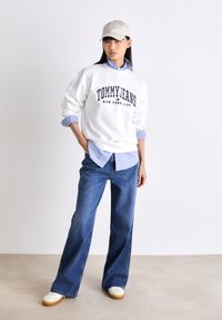 Sudadera blanca con el texto "TOMMY JEANS NEW YORK CITY", combinada con una camisa de color azul claro, pantalones vaqueros de pierna ancha en azul y zapatillas blancas.