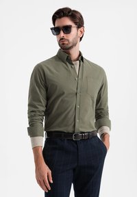 Olijfgroen button-up shirt met een borstzak, gedragen over een lichtgekleurde ronde hals, gecombineerd met marineblauwe geruite broek en een zonnebril.