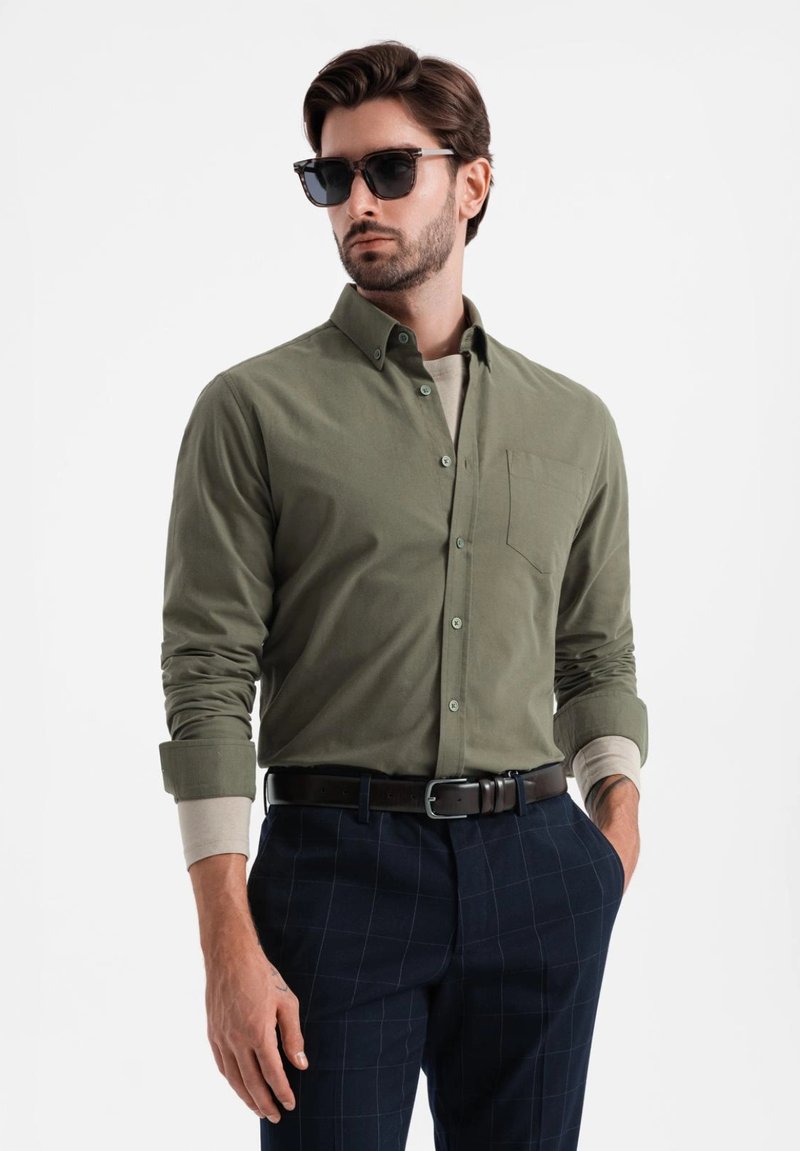 Olijfgroen button-up shirt met een borstzak, gedragen over een lichtgekleurde ronde hals, gecombineerd met marineblauwe geruite broek en een zonnebril.