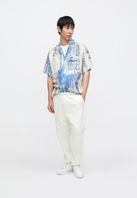 Koszula z krótkim rękawem z niebieskim i białym abstrakcyjnym wzorem; kremowe plisowane spodnie i białe sneakersy. Prosty fason o luźnym kroju.