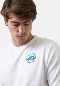 Camiseta de manga larga blanca con un logo gráfico azul claro en la parte izquierda del pecho, cuello redondo y textura de algodón suave.