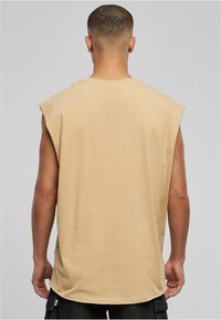 Beige mouwloos shirt met een ontspannen pasvorm, zachte katoenen stof, afgeronde zoom en minimale stikseldetails. Achteraanzicht toont een gladde textuur.