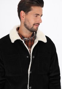 Sort fløjljakke med hvid faux-shearling krave, der har to frontlommer og trykknapper. Båret over en plaidskjorte.