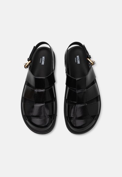 Black leather sandals with woven design, open back, and ankle strap featuring a gold-colored hardware detail. Smooth, glossy texture.