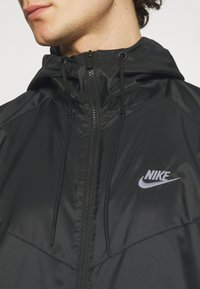 Černá bunda Nike s předním zipem, nastavitelnou kapucí a logem na hrudi. Hladký, lehký materiál s přiléhavým střihem.