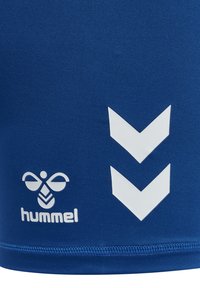 Gros plan sur un tissu bleu avec un logo d'abeille hummel blanc et deux chevrons orientés vers le bas imprimés côte à côte près de l'ourlet cousu.