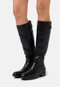 Černé kožené kozačky ke kolenům s nízkým podpatkem, bočním zipem a ozdobnou přezkou, nošené na bosých nohách na bílém pozadí.