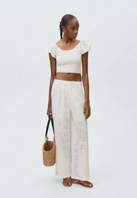 Top din bumbac alb, scurt, cu mâneci volan, asortat cu pantaloni largi cu model de perforație. Modelul ține o geantă din fibră naturală și poartă sandale.