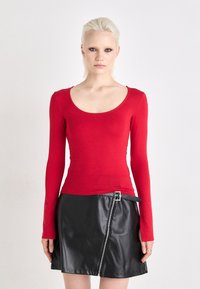Rotes Langarmoberteil mit weitem Halsausschnitt, kombiniert mit einem schwarzen Kunstlederrock, der einen seitlichen Reißverschluss und Gürtelschlaufen aufweist.