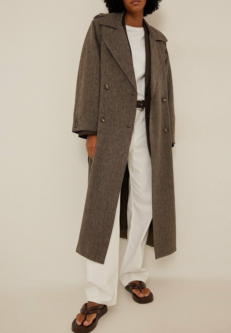 Personne portant un long manteau marron à double boutonnage sur une chemise blanche, un pantalon blanc à larges jambes et des sandales plateformes marron foncé.