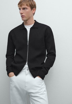 Giovane uomo con le mani in tasca, che indossa un maglione nero con zip sopra una camicia bianca e pantaloni bianchi, che guarda di lato su uno sfondo uniforme.