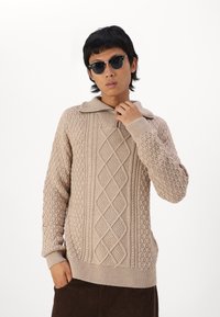 Pull en tricot beige avec un col montant, des motifs texturés et une fermeture zippée. Modèle portant des lunettes de soleil noires, les mains sur le col.
