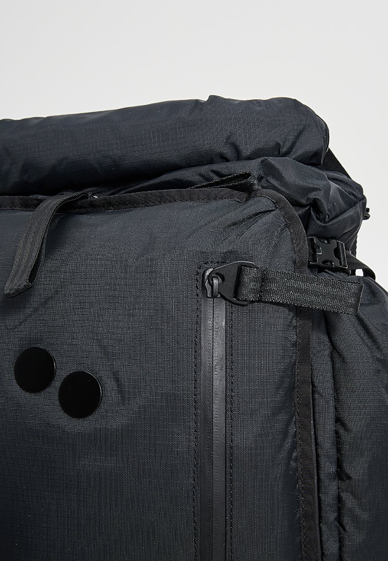 Schwarzer Stoffrucksack mit Rollverschluss, seitlicher Reißverschlusstasche, verstellbarem Riemen und zwei runden schwarzen Aufnähern auf der Vorderseite.
