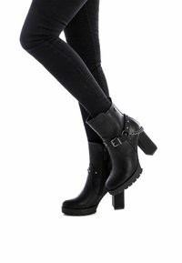 Botas de tobillo negras con tacón, hechas de material sintético, que cuentan con un cierre lateral, detalles metálicos y un tacón grueso con una suela robusta.