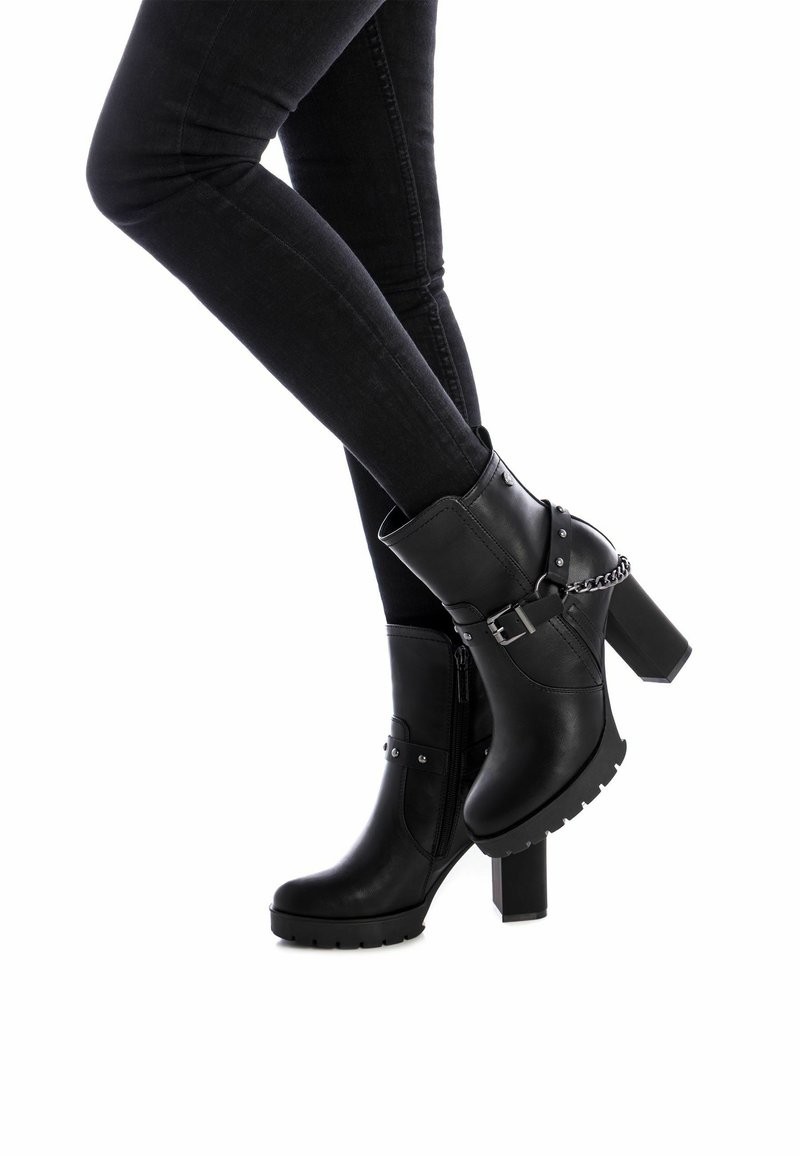 Botas de tobillo negras con tacón, hechas de material sintético, que cuentan con un cierre lateral, detalles metálicos y un tacón grueso con una suela robusta.